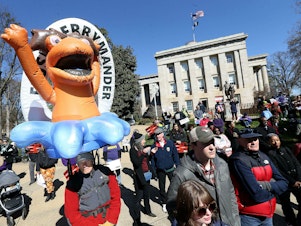 caption: Demonstrators gather outside the North Carolina State Capitol in Raleigh, N.C., with a "FLUSH GERRYMANDER" inflatable on the day the state's highest court held a rehearing for the redistricting case known nationally as <em>Moore v. Harper</em>.