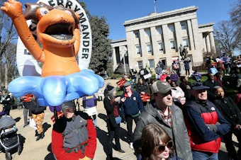 caption: Demonstrators gather outside the North Carolina State Capitol in Raleigh, N.C., with a "FLUSH GERRYMANDER" inflatable on the day the state's highest court held a rehearing for the redistricting case known nationally as <em>Moore v. Harper</em>.