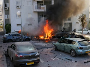 caption: A member of the Israeli security forces stands near burning cars following a rocket attack from the Gaza Strip in Ashkelon, southern Israel, on Oct. 7.