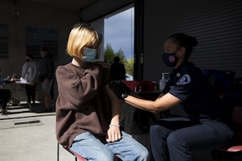 caption: Clem Watts, a 17-year-old junior at The Center School, receives a Pfizer Covid-19 vaccine, administered by Seattle Fire Captain Melissa Woolsey, right, on Tuesday, May 18, 2021, at Memorial Stadium in Seattle. 