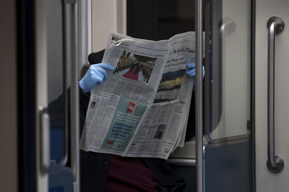 caption: A light rail passenger reads the Seattle Times while wearing blue gloves on Thursday, March 26, 2020, at Westlake Station in Seattle. 