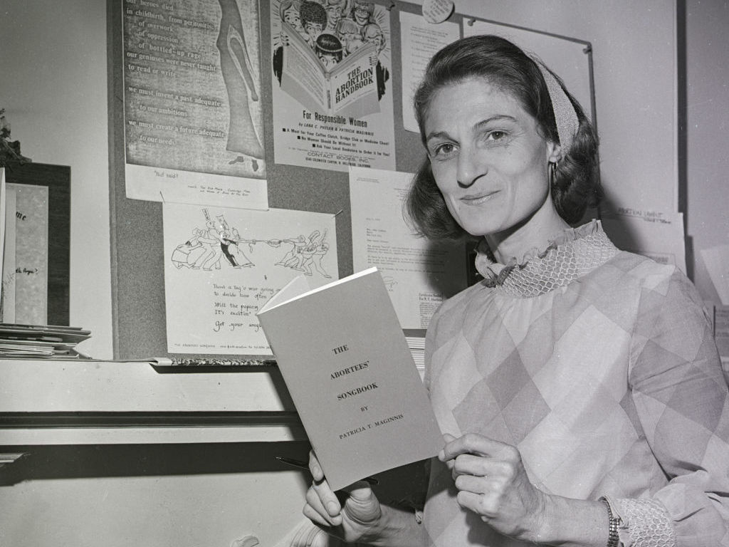 caption: Patricia Maginnis holds her <em>Abortees' Songbook</em> and stands next to a bulletin board full of abortion information. The early abortion rights advocate, pictured here in 1970, died earlier this year.