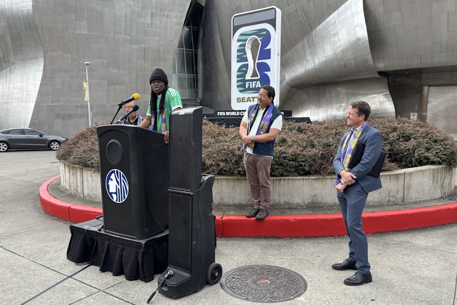 caption: Sounders FC owner and former player Steve Zakuani speaks as Mayor Bruce Harrell (center) and Seattle Center Director Marshall Foster look on.
