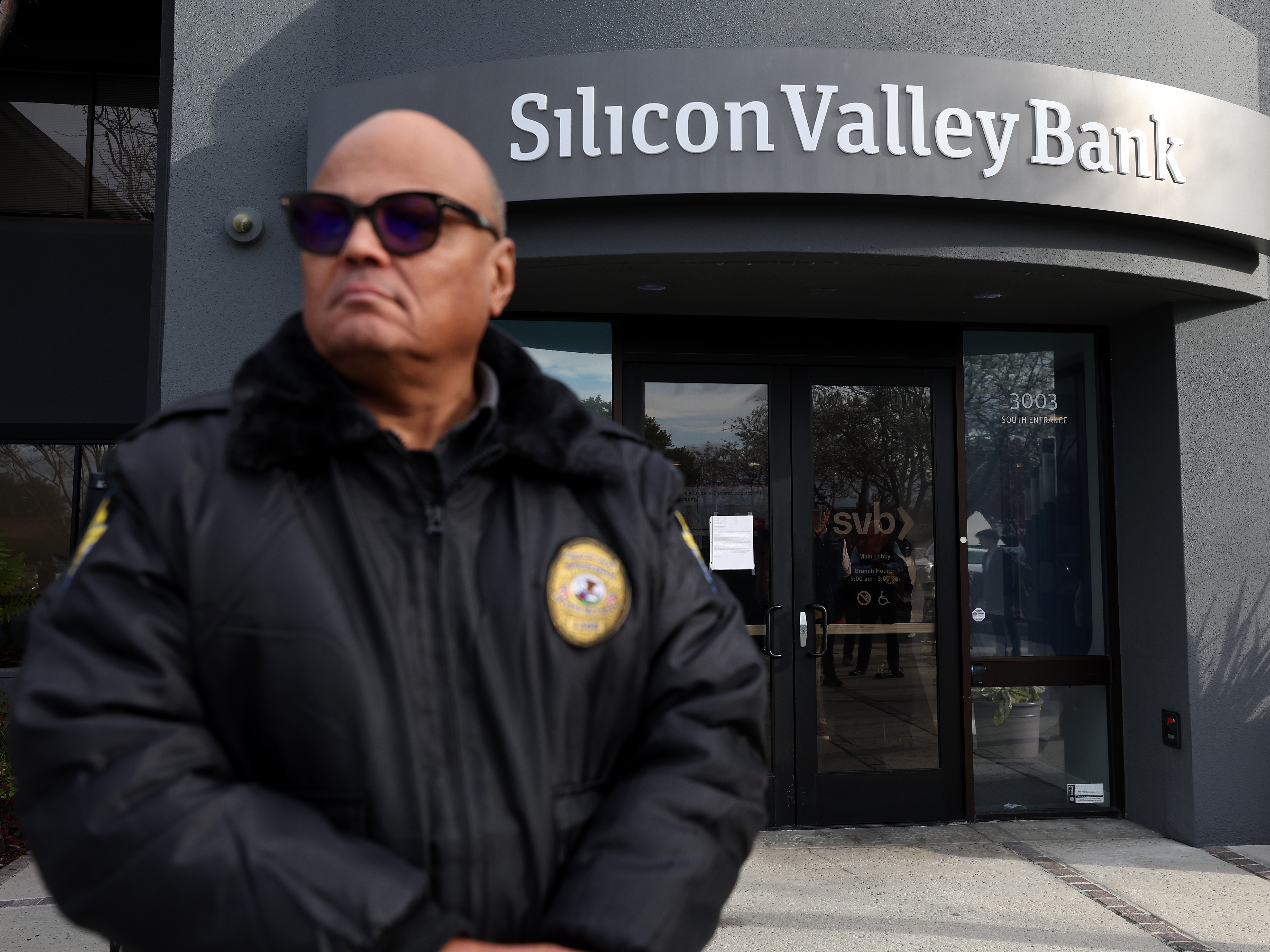caption: A security guard at Silicon Valley Bank monitors a line of people outside the office in Santa Clara, Calif., on March 13, 2023. The Fed admitted it was partly to blame for the collapse of the lender in a scathing report on Friday.