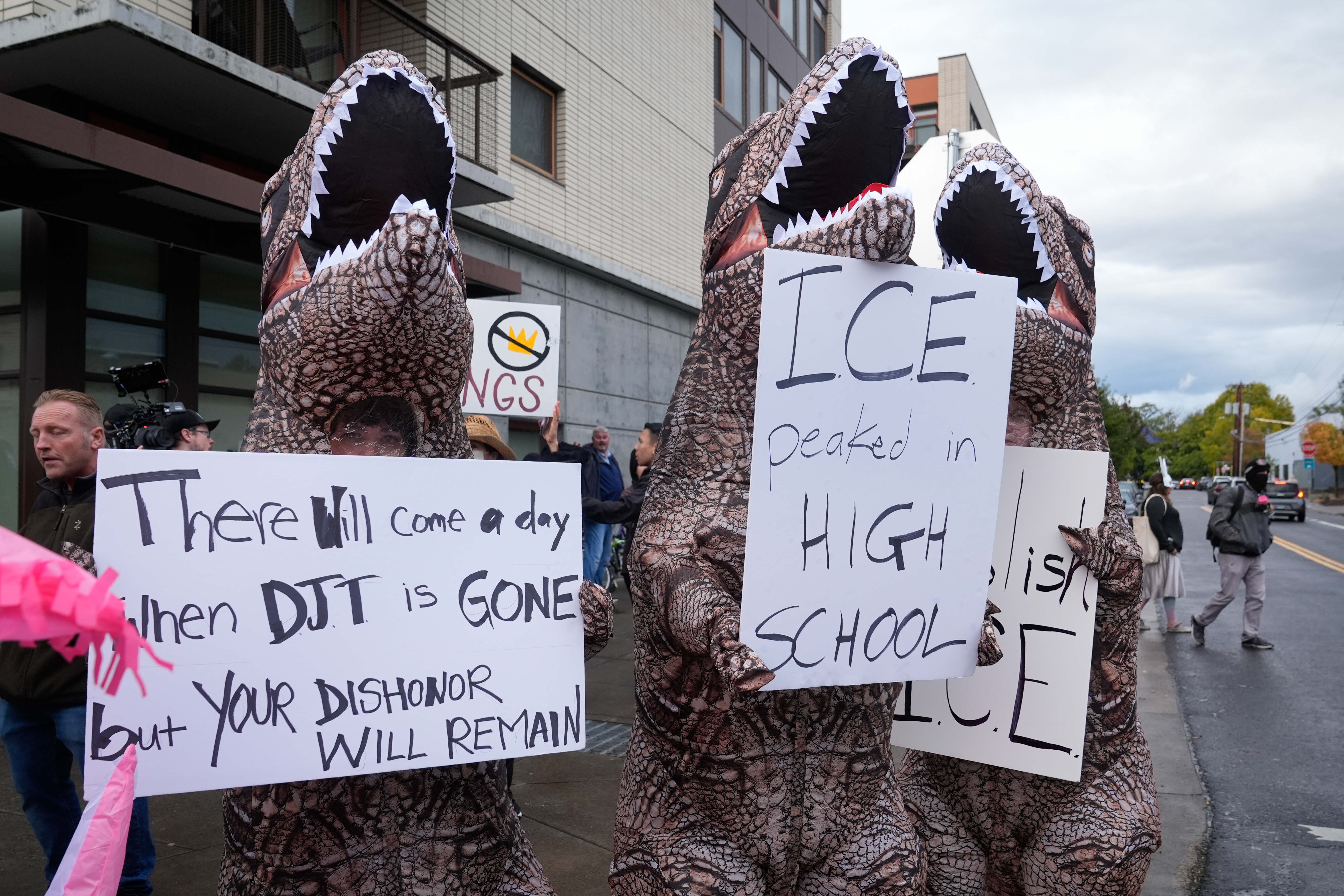caption: Demonstrators in inflatable dinosaur costumes hold signs in protest against Donald Trump's immigration policies, Sunday, Oct. 12, 2025, in Portland, Oregon. 