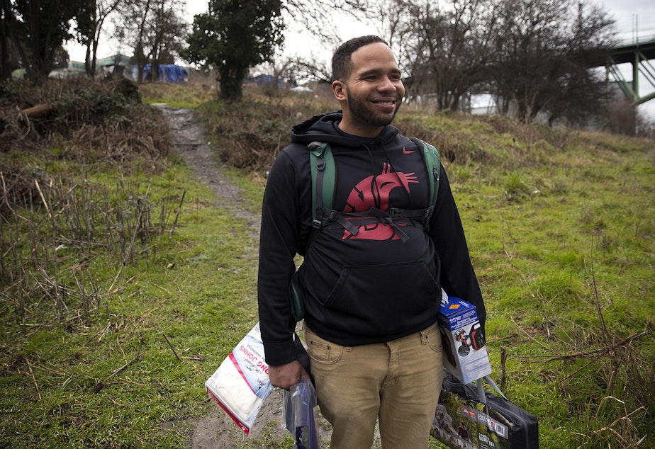caption: Kalamante Fleeks, 29, smiles while holding mutual aid supplies including long johns, a tarp, a new tent, and a portable power pack delivered to him by Joscelyn and Cass DuVani on Friday, March 5, 2021, in Seattle. 