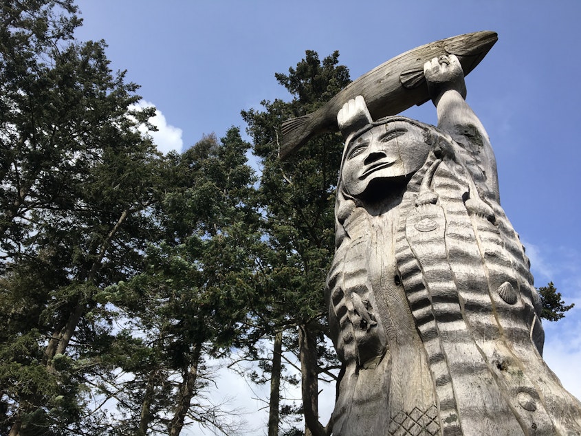 caption: A cedar carving of Ko-Kwal-Alwoot, the Maiden of Deception Pass, with hair turned to ribbons of kelp, holds a salmon aloft at Deception Pass State Park.