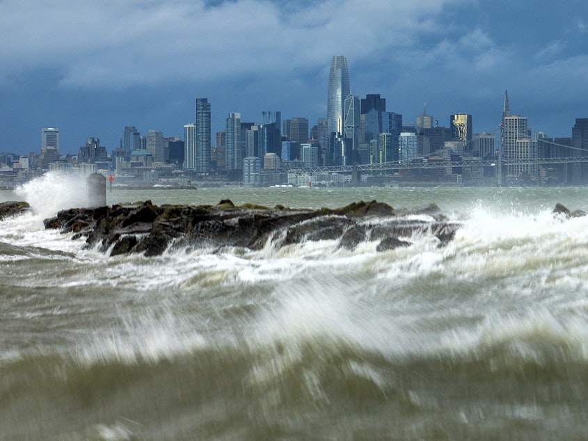 caption: Waves crash over a breakwater in Alameda, Calif., with the San Francisco skyline in the background on Sunday, Feb. 4, 2024.