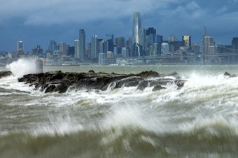 caption: Waves crash over a breakwater in Alameda, Calif., with the San Francisco skyline in the background on Sunday, Feb. 4, 2024.