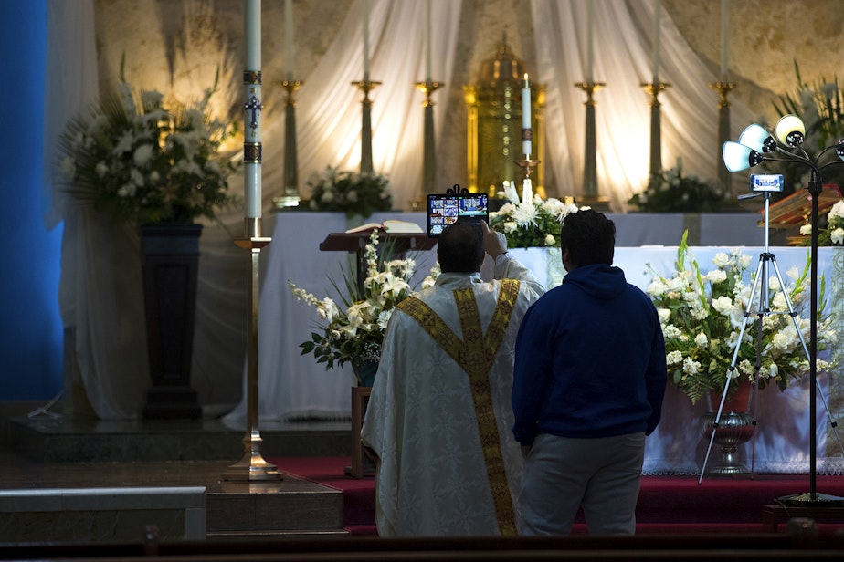 caption: Father Jose Alvarez has help setting up an iPad ahead of a live-streamed daily mass on Friday, April 24, 2020, at Holy Family Roman Catholic Church in White Center. 