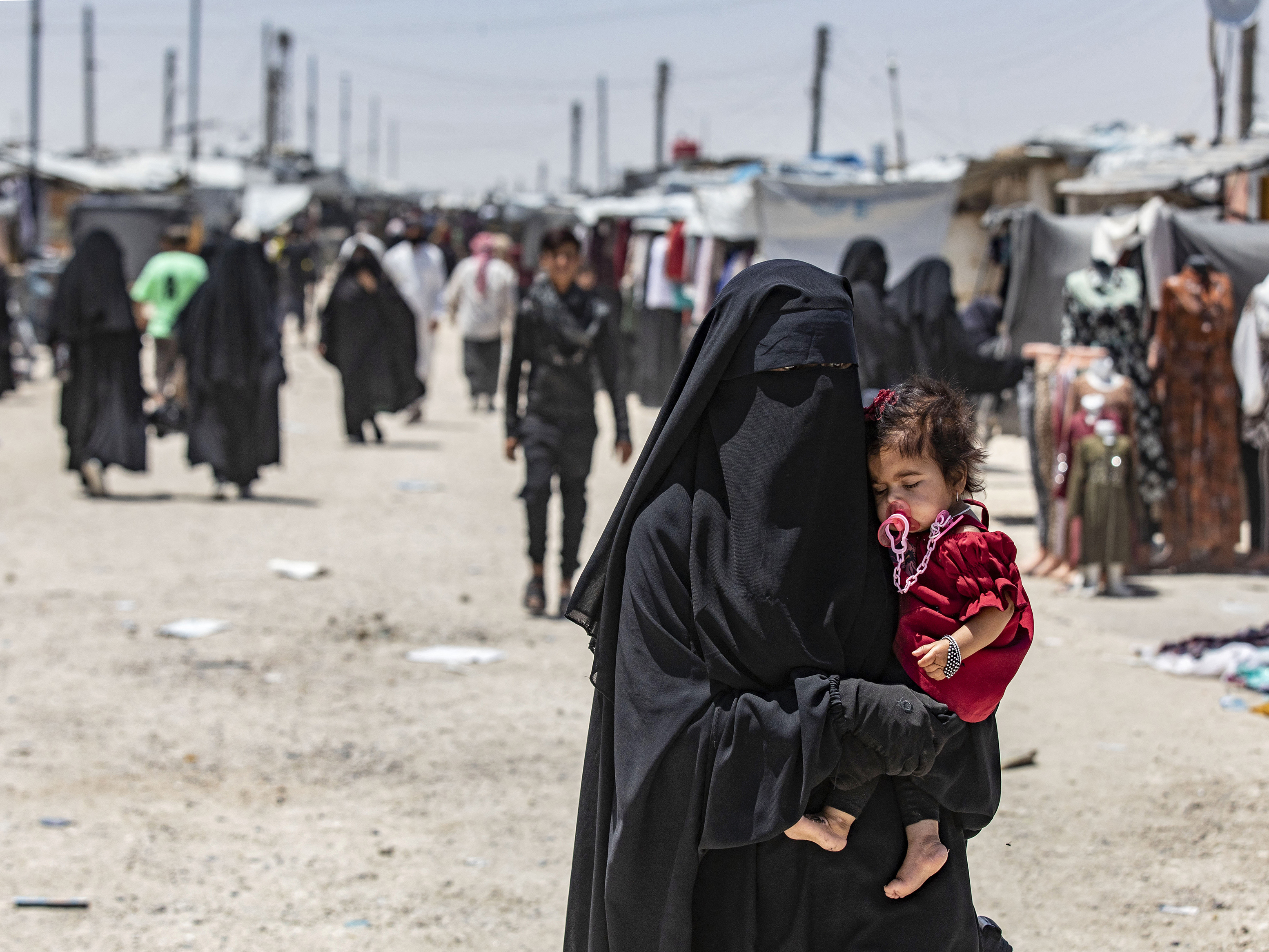 caption: A woman carries a child as she walks through the al-Hol refugee camp in northeastern Syria in Oct. 2023.