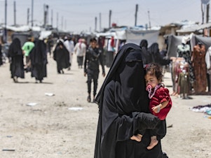 caption: A woman carries a child as she walks through the al-Hol refugee camp in northeastern Syria in Oct. 2023.