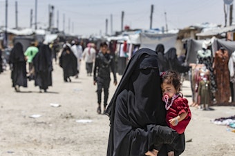 caption: A woman carries a child as she walks through the al-Hol refugee camp in northeastern Syria in Oct. 2023.