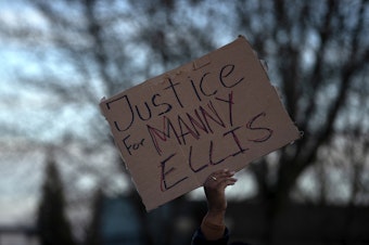caption: Justice for Manny Ellis reads a handwritten sign as a crowd gathered at the intersection of 11th Street and Martin Luther King Jr. Way, after a jury found three Tacoma police officers not guilty in the trial over the death of 33-year-old Manuel Ellis, on Thursday, December 21, 2023, in Tacoma. 
