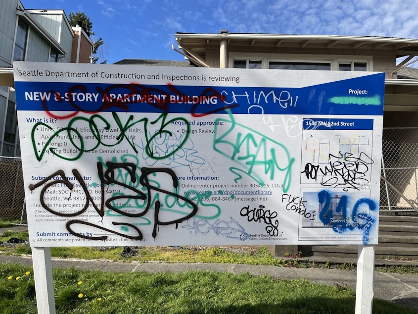 caption: A vandalized sign in front of a the construction site for a dense apartment building with small apartment units (by another developer).
