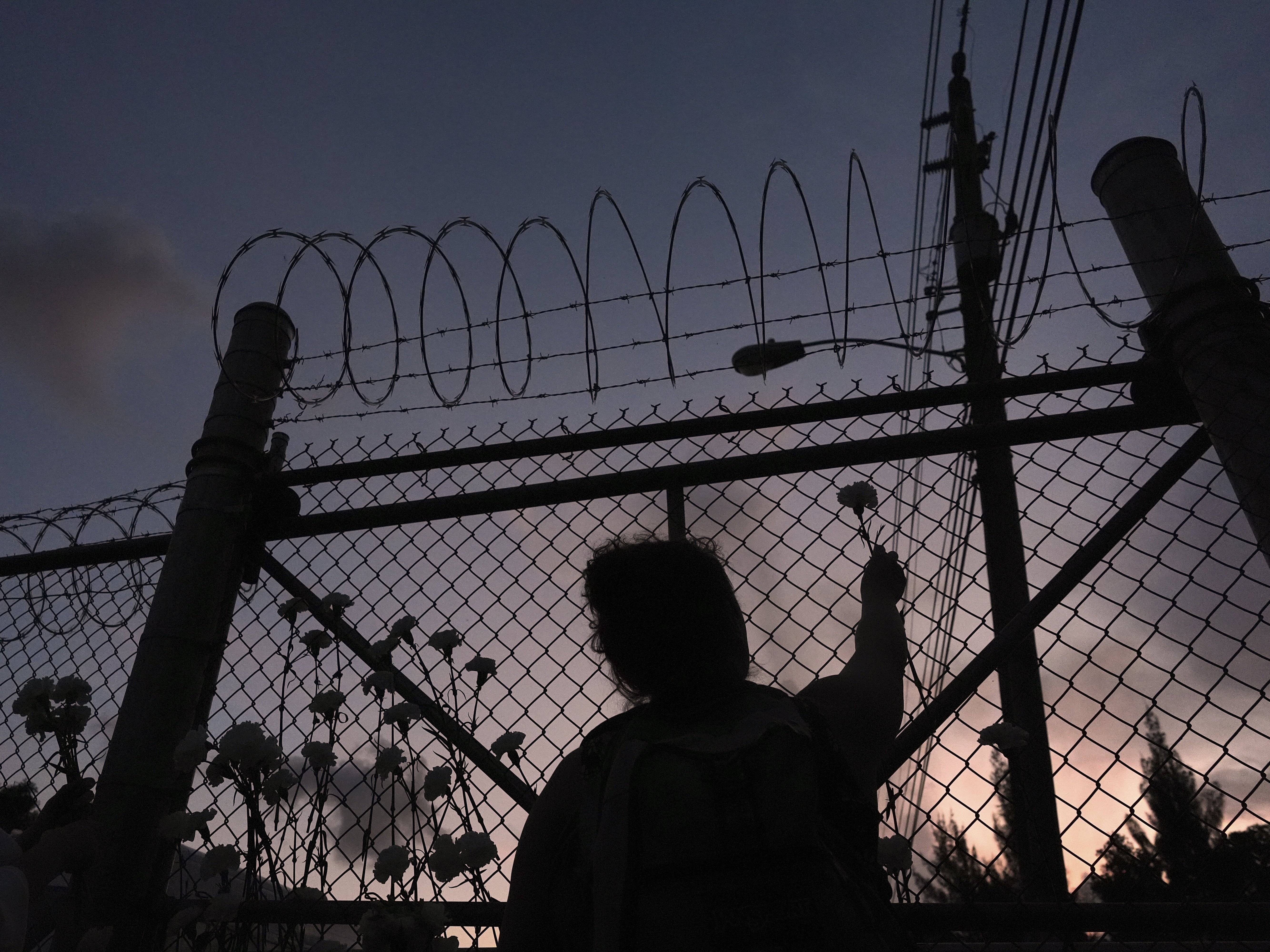 caption: People place flowers on a fence outside Krome Detention Center in Miami in May 2025, during a vigil to recognize people who have died in U.S. Immigration and Customs Enforcement custody as well as those affected by mass deportations.