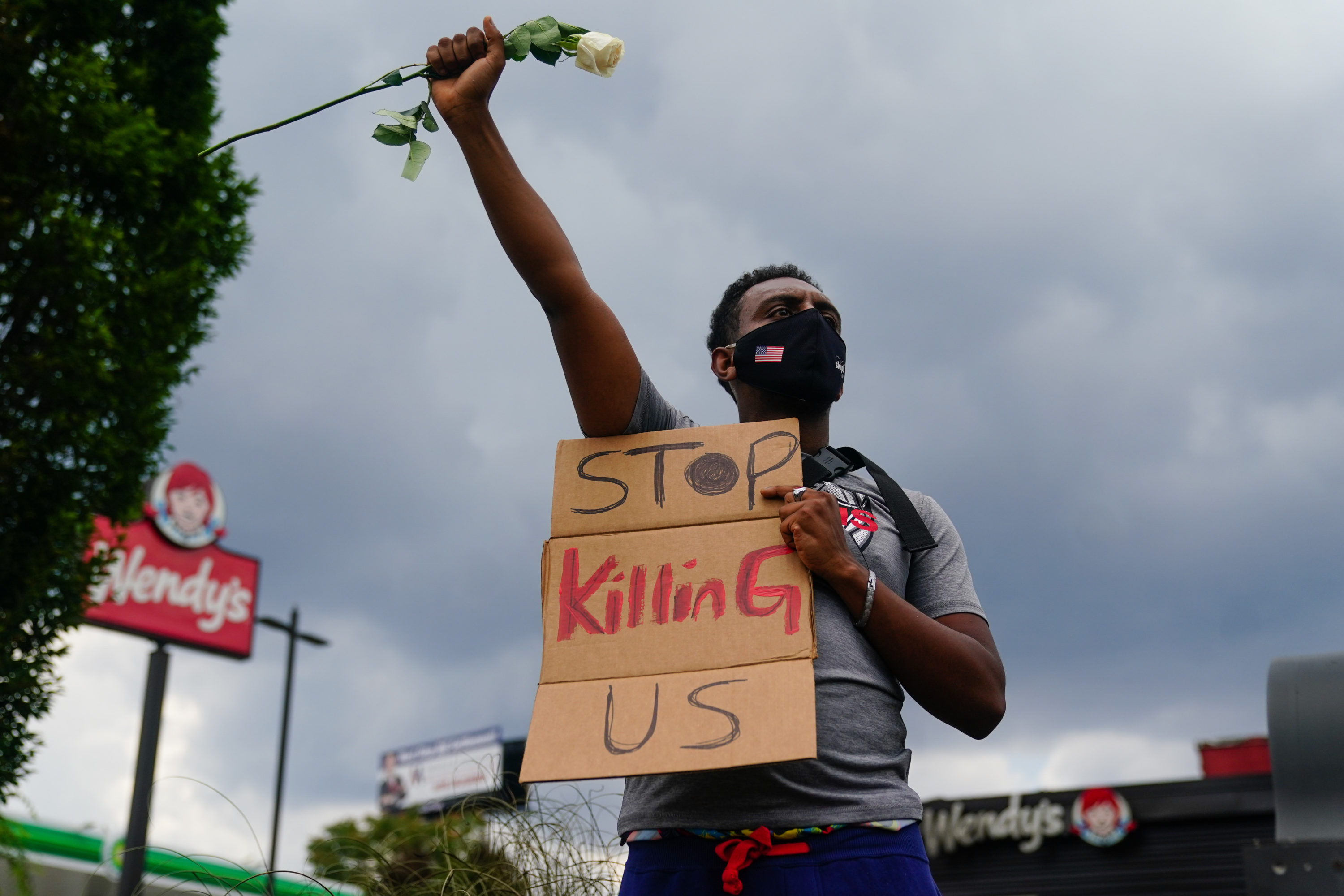 caption: A man holds a sign and a white rose in his fist while facing traffic outside a burned Wendy's restaurant on the second day following the police shooting death of Rayshard Brooks in the restaurant parking lot June 14, 2020, in Atlanta, Georgia. (ELIJAH NOUVELAGE/AFP via Getty Images)