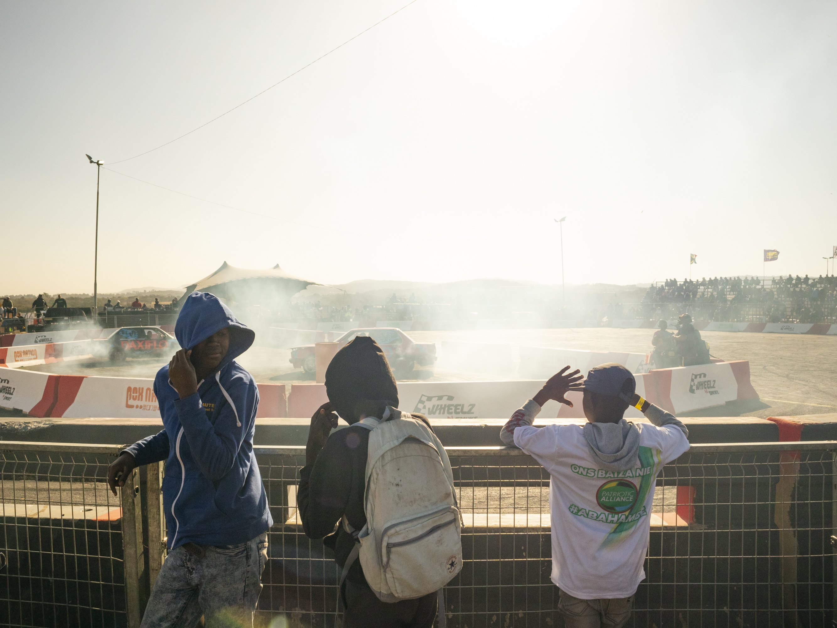 caption: Three young spectators cover their faces from rubber debris and smoke at the spinning field, at Wheelz N Smoke arena, on July 7, in Johannesburg, South Africa.