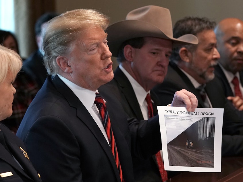 caption: President Trump holds a picture labeled "typical standard wall design" as he hosts a roundtable discussion on border security in the Cabinet Room of the White House on Friday.