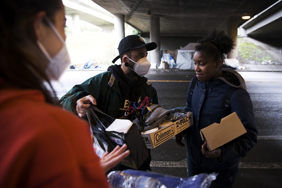caption: From left, Joscelyn and Cass DuVani deliver a new tent, shoes, clothing, soup, and a tarp, along with several other items to Tedra, 24, who is currently experiencing houselessness, on Friday, March 5, 2021, underneath Interstate 5 along South Dearborn Street in Seattle. Tedra's tent and belongings had been damaged after catching fire the previous evening. KUOW Photo/Megan Farmer 