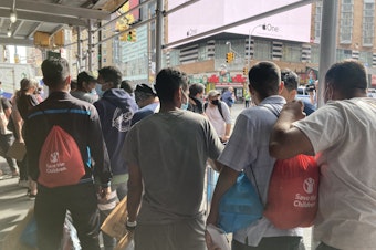 caption: Asylum-seekers who have recently arrived in New York on a bus from Texas wait for ride-share vehicles, organized by a mutual aid group, to take them into the city.