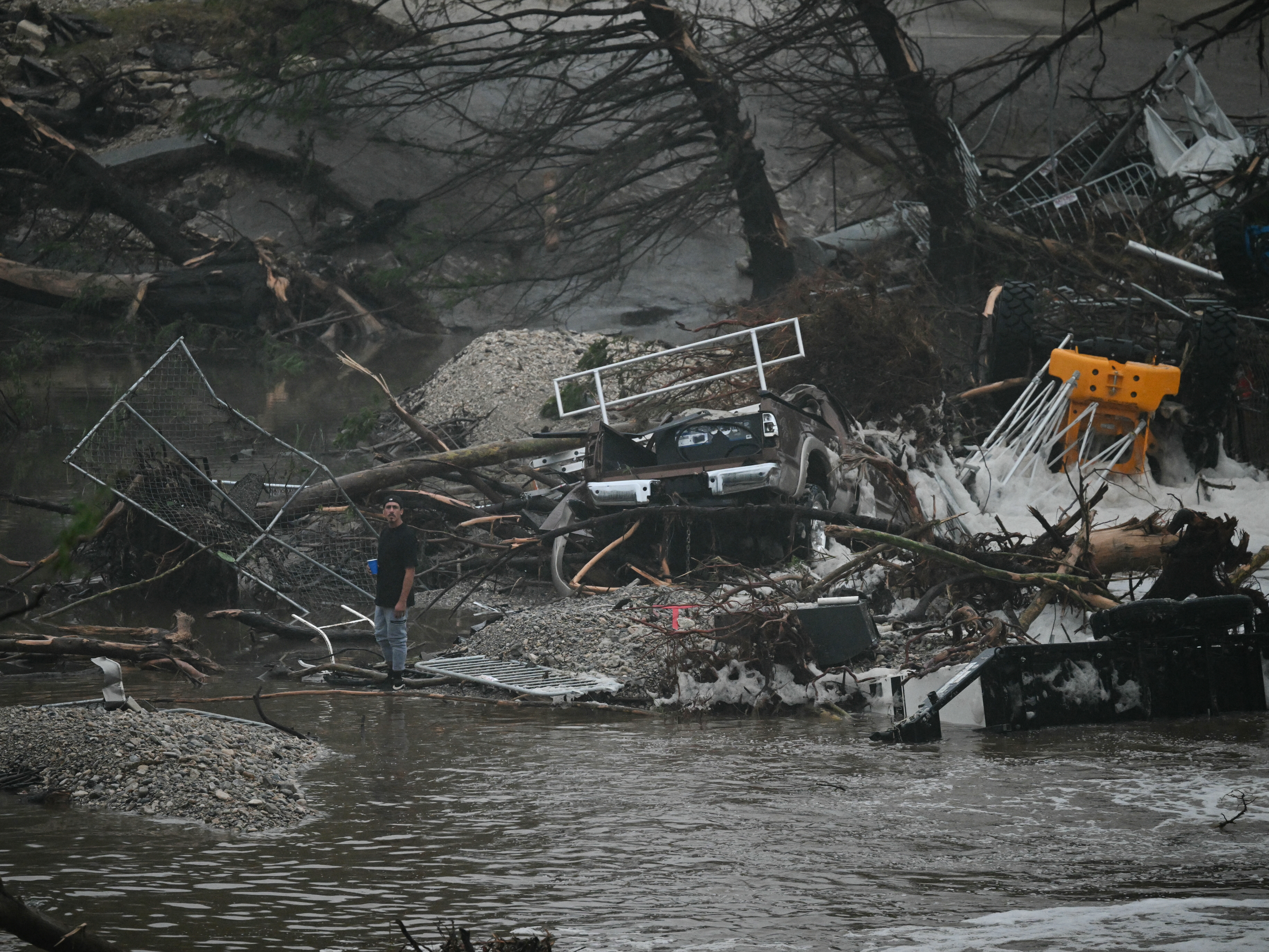 caption: A member of the public stands next to overturned vehicles and broken trees on Saturday after a flash flood at the Guadalupe River in Kerrville, Texas.