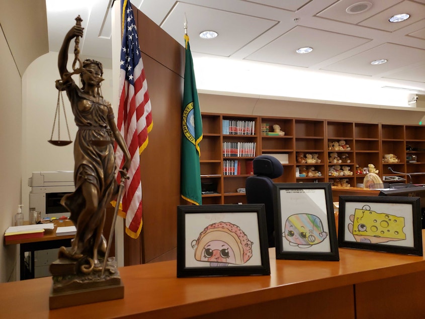 caption: Inside Judge Rajul's court room, Lady Justice stands next to framed coloring pages she's received from migrant children. In the background, rows of teddy bears wait to support youth during the long immigration process. 
