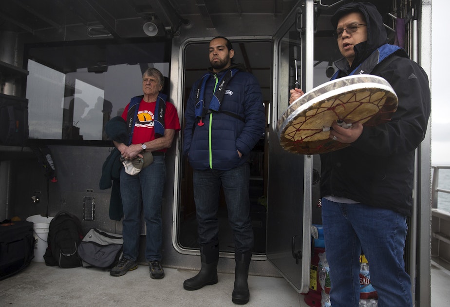 caption: From left, Kurt Russo and Tony Hillaire listen as Lawrence Solomon, right, sings the Lummi National Anthem before a ceremonial feeding where one live chinook salmon was released into the water, aboard King County Research Vessel SoundGuardian on Wednesday, April 10, 2019, near Henry Island.