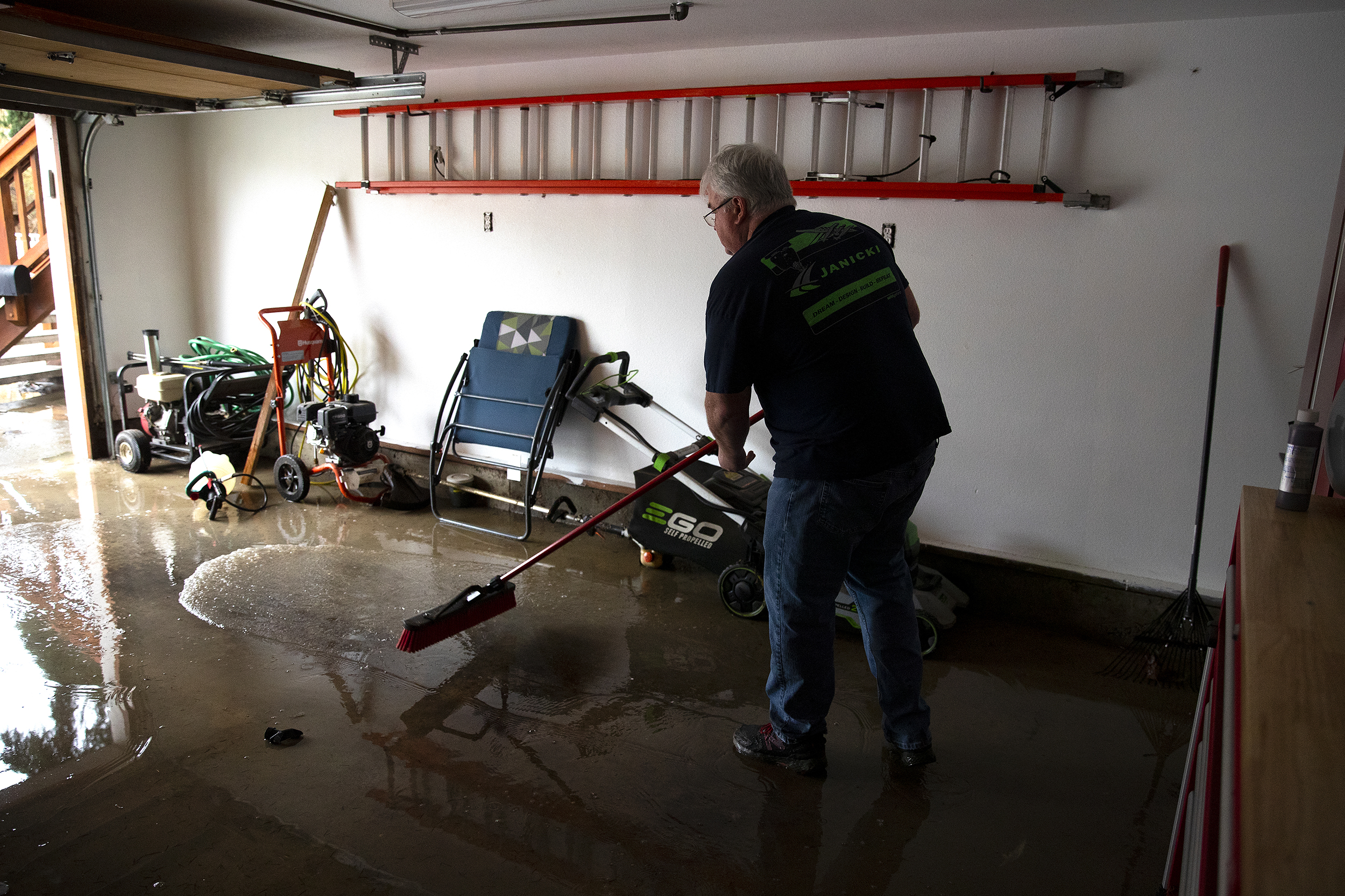 caption: Mike Leander pushes flood water out of his garage with a large broom on Friday, December 12, 2025, in Burlington. 