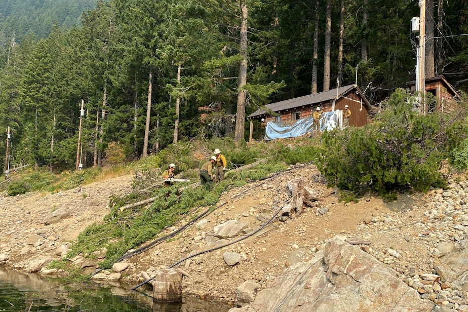 caption: Firefighters wrap a cabin at Ross Lake Resort with aluminum-fiberglass foil for fire protection on Aug. 7, 2023.