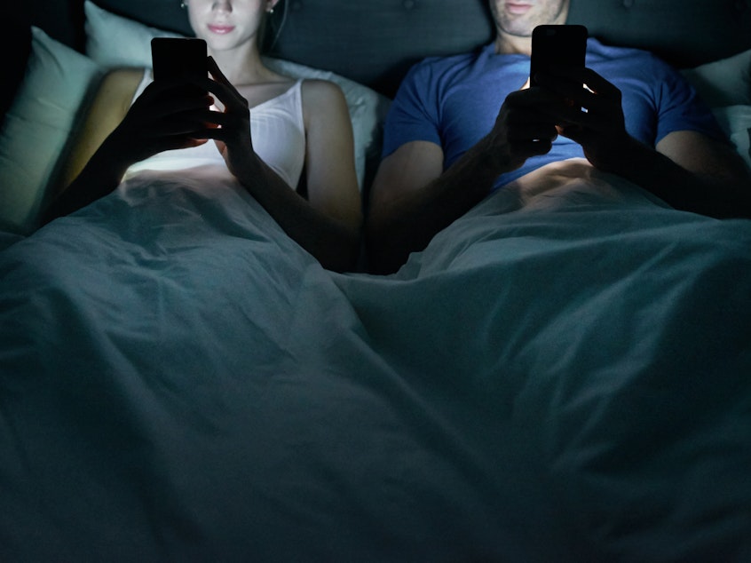 A couple side by side in bed, each looking at their cell phones.