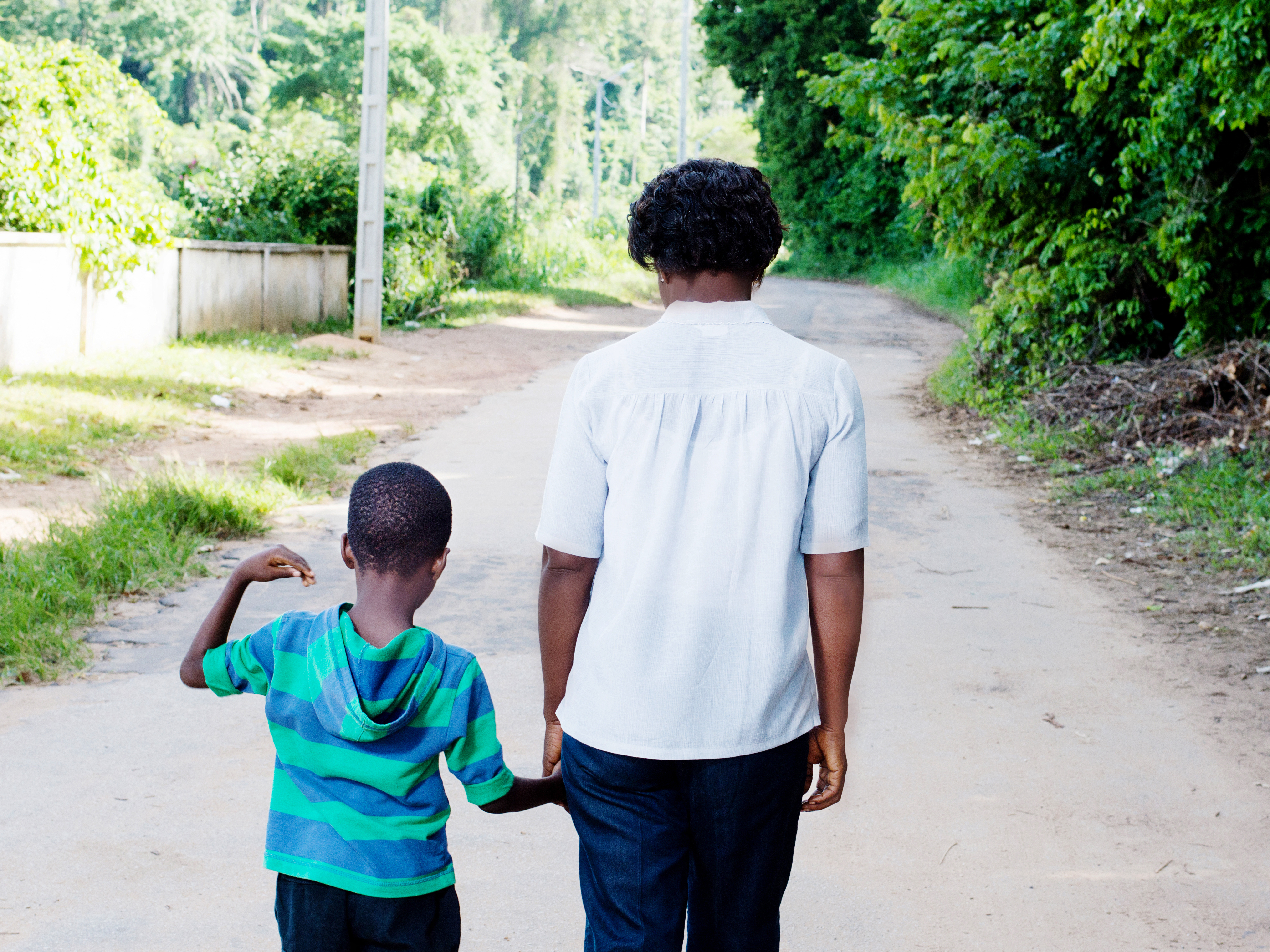 A mother and son walk hand in hand down a pathway, facing away from the camera.