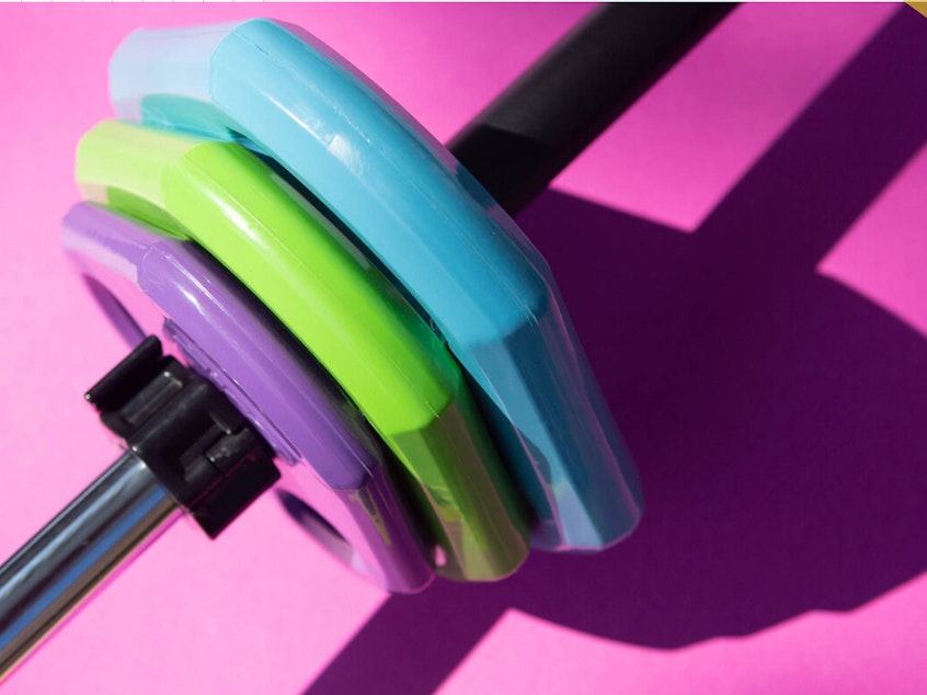 Close up photo of a barbell with three colorful weights on the end against a pink background.