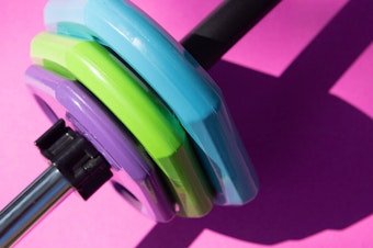 Close up photo of a barbell with three colorful weights on the end against a pink background.