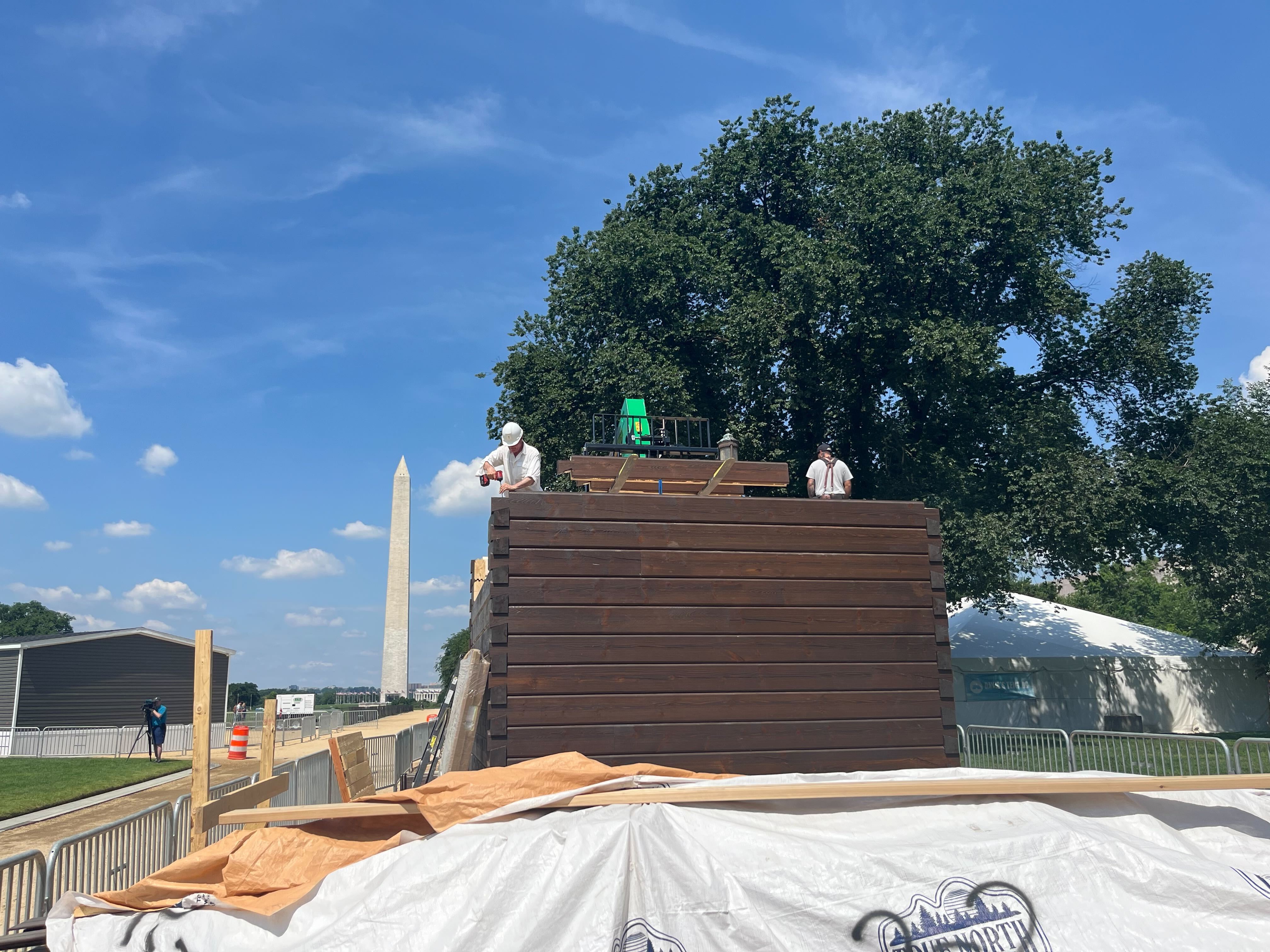 caption: Workers assemble a home resembling an instant log cabin on the National Mall in Washington, D.C., with the Washington Monument behind them.