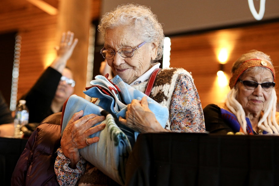 caption: Lummi elder Ernestine Lane receives a gift after sharing her experiences at the Chemawa Indian School in Salem, Oregon. Interior Secretary Deb Haaland  visited the Tulalip Gathering Hall on Sunday, April 23, 2023, during the “Road to Healing” tour. The yearlong tour provides opportunity for Indigenous survivors of the federal Indian boarding school system to share their experiences. 