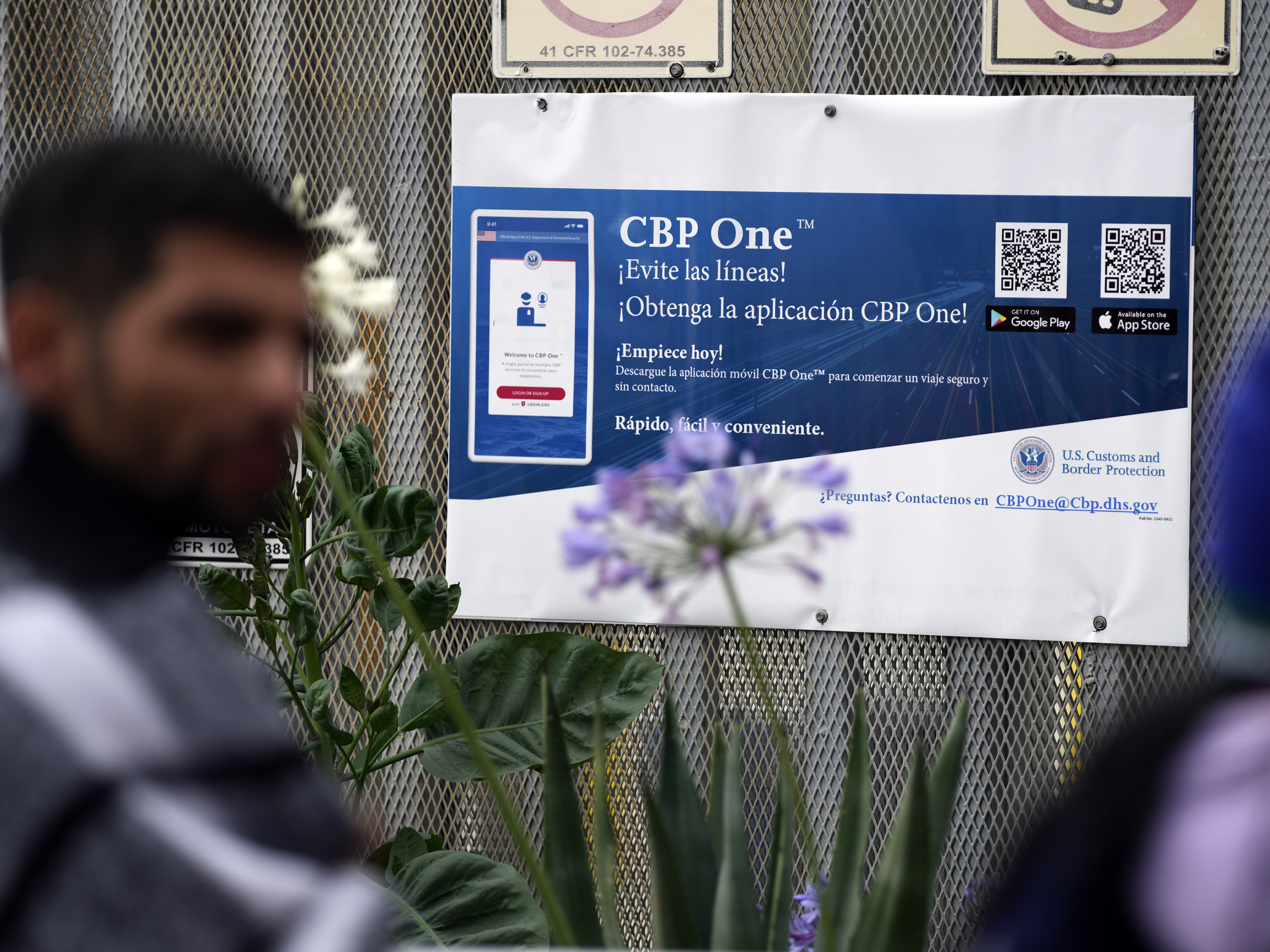 caption: People waiting to apply for asylum stand in front of a sign for the CBP One app as they camp near the pedestrian entrance to the San Isidro Port of Entry, linking Tijuana, Mexico with San Diego.