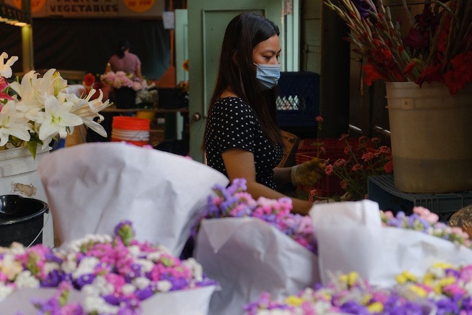 caption: Kim Cruz works on a bouquet at Santos Farmers flower booth at Pike Place Market, August 2, 2021. Santos Farmers owner, Eric Santos, says business has still not picked back up enough since 2020 and they are struggling to make money. Pre-pandemic business would be good all week during the summer, now he can only count on weekend days to be profitable. He hopes local people will continue to support the small business owners at the market.