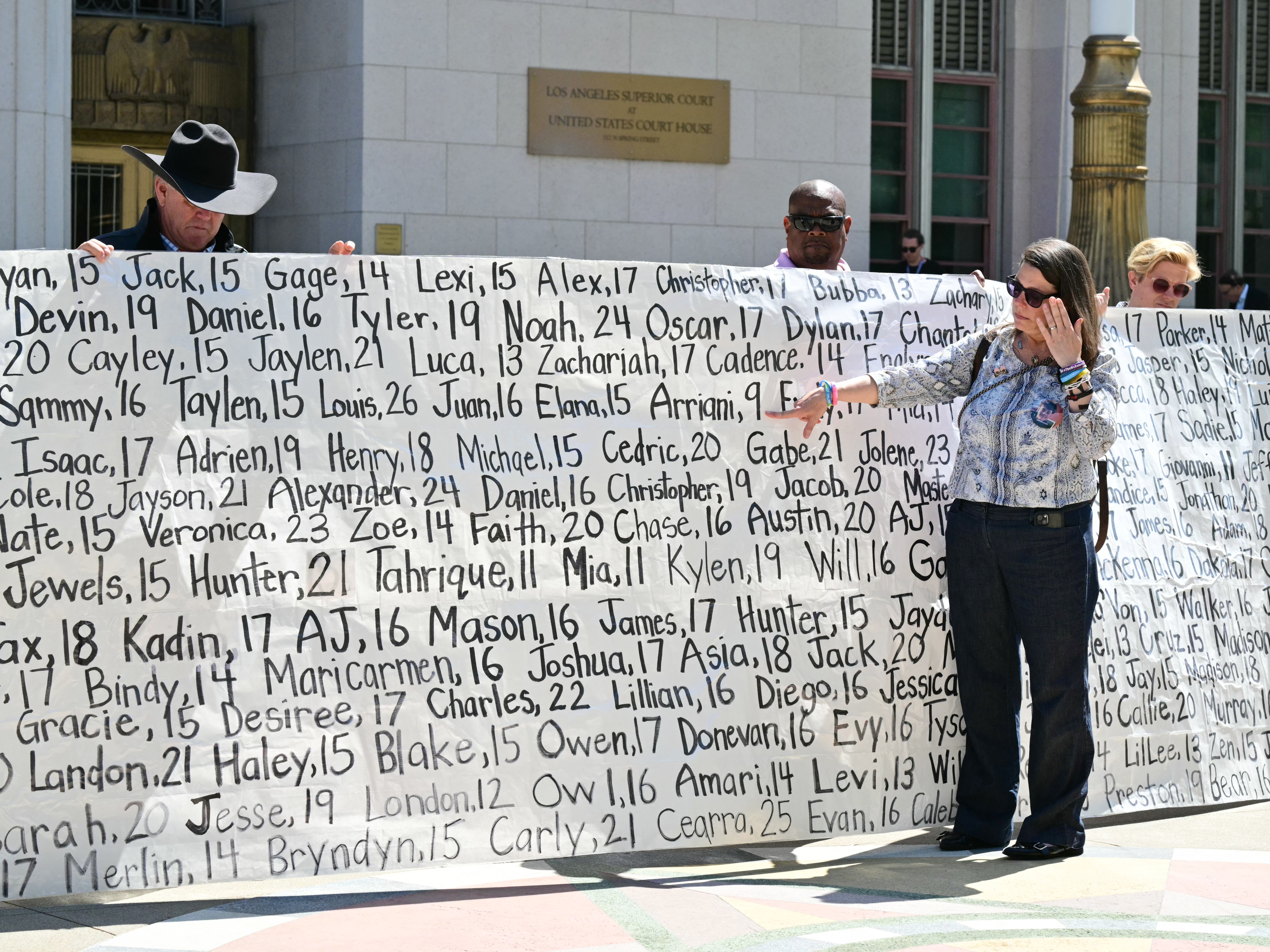 caption: Mary Rodee, whose 15-year-old son died by suicide, points to a banner listing victims' names outside Los Angeles Superior Court on March 25, 2026, after a jury found Meta and YouTube liable for harming a young woman through the addictive design of their social media platforms. The landmark verdict could reshape how the tech industry faces legal accountability for harms to users.
