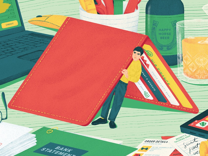 Illustration of a person in miniature hiding underneath a tented wallet on a desk, peering out to a tablescape of bills, finance apps, a laptop, receipts and a few drinks, symbolizing hiding from money problems.