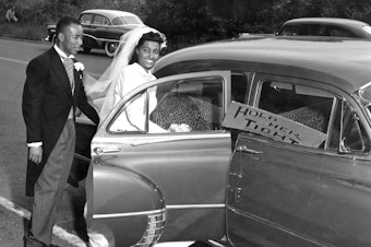 caption: Newlywed bride and groom stepping into car, circa 1955.  Sign in front passenger side window reads "Hold Her Tight."