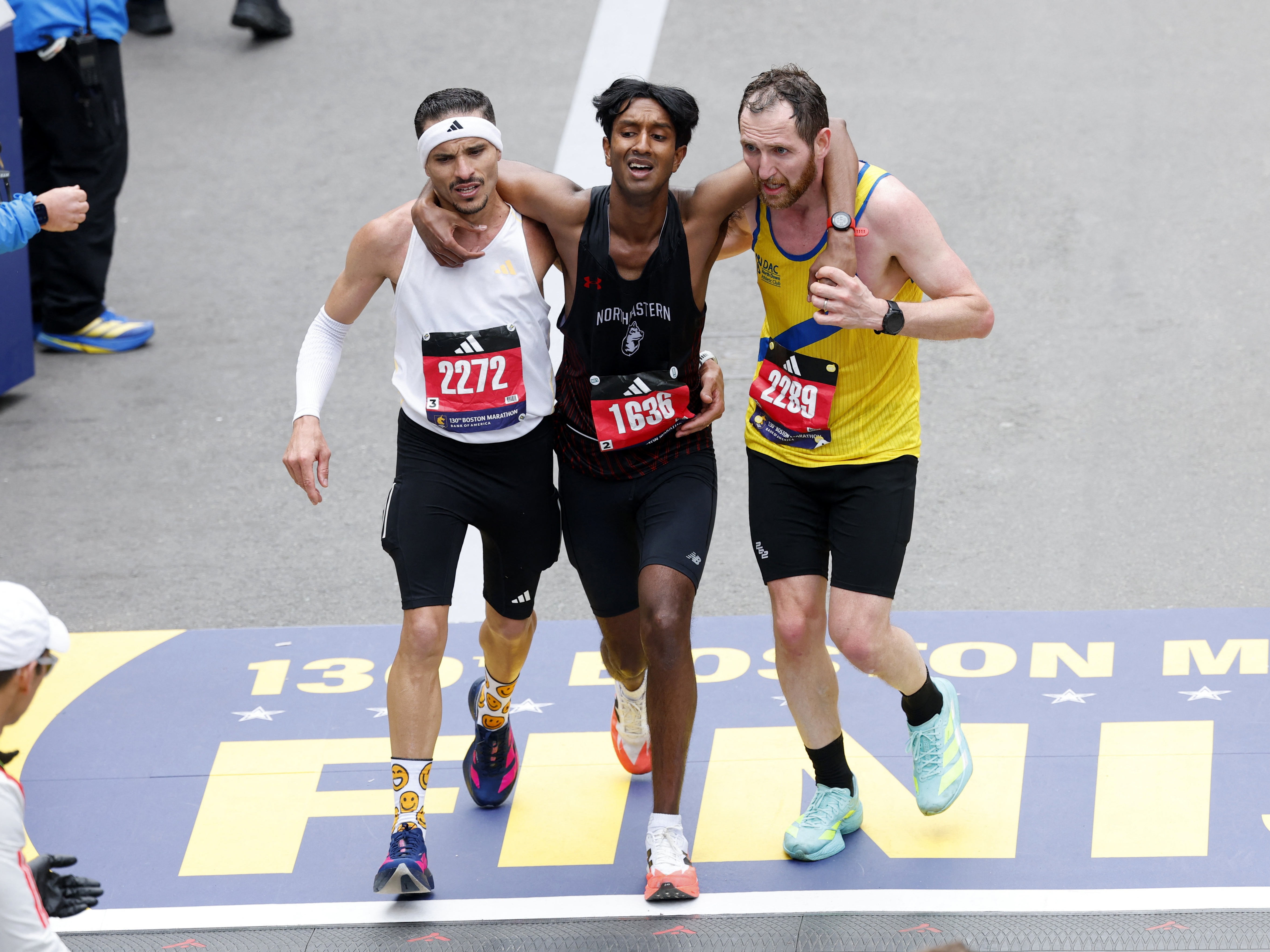 caption: Robson De Oliveira, Ajay Haridasse and Aaron Beggs cross the finish line in the Boston Marathon on Monday.