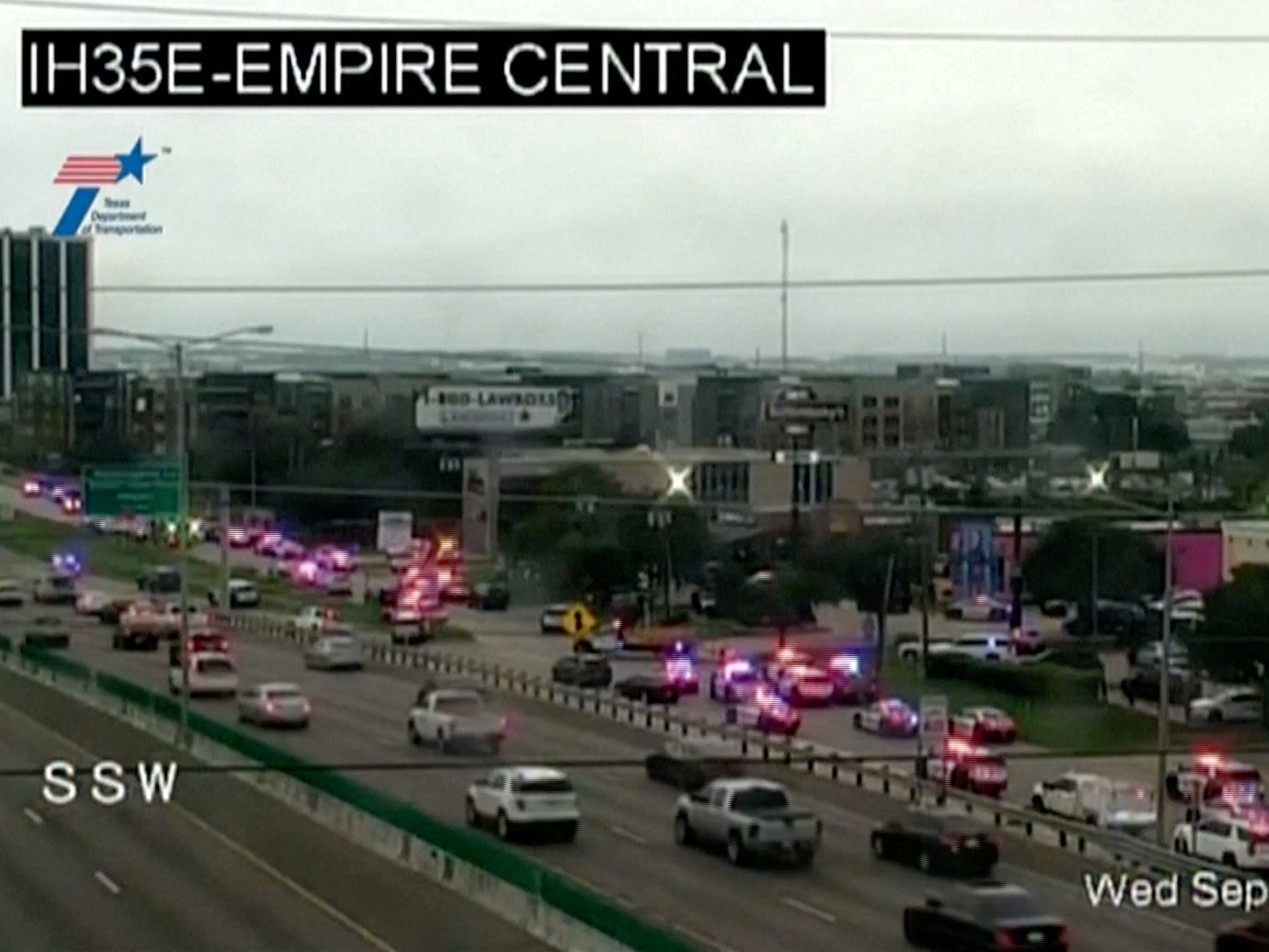 caption: Police cars and ambulances wait outside an Immigration and Customs Enforcement detention facility in Dallas where a shooting took place on Wednesday morning, in this screenshot from a Texas Department of Transportation traffic camera video.