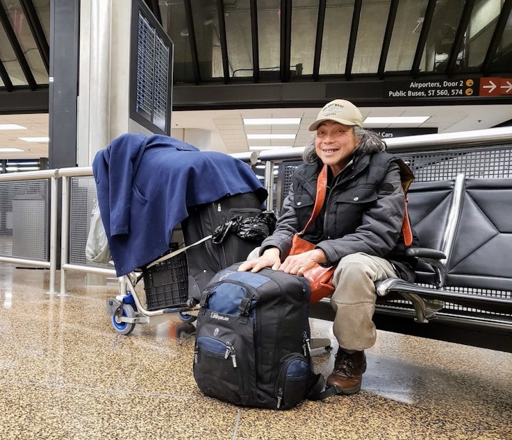 caption: De Chung sits with his bags and belongings near a luggage carousel at SeaTac Airport on Tuesday, Jan. 3, 2023.