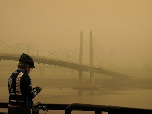 caption: A man stops on his bike along the Willamette River as smoke from wildfires partially obscures the Tilikum Crossing Bridge in Portland, Ore., Saturday.