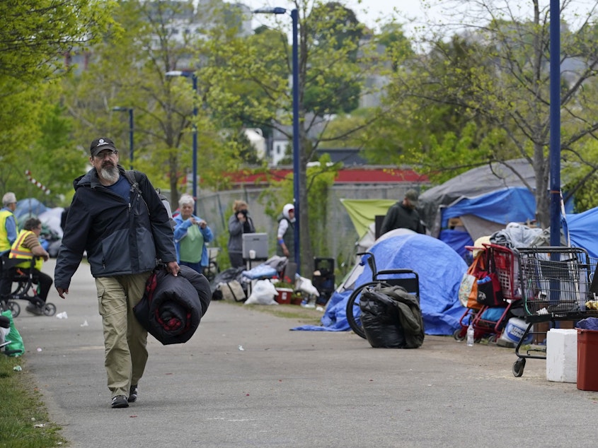 caption: A man carries a sleeping bag at a homeless encampment in Portland, Maine, in May, before city workers arrived to clean the area. State officials say a lack of affordable housing is behind a sharp rise in chronic homelessness.