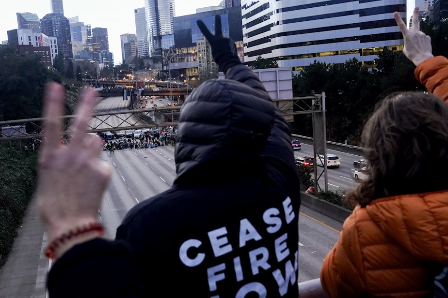 caption: Protesters calling for a cease-fire in the Israel-Hamas war gesture a peace sign on an overpass to those below as they block Interstate 5 northbound Saturday, Jan. 6, 2024, in downtown Seattle. 