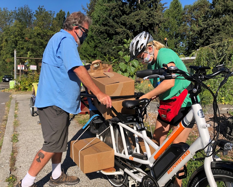 caption: Paul Dieter steadies the bike while Ellen Nichter secures the load that will be delivered to families nearby. Dieter and Nichter are bike volunteers helping food banks with home deliveries. 