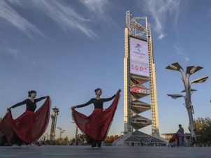 caption: Women practice a dance routine on Oct. 27 in front of a large countdown screen showing 100 days before the opening of the Beijing 2022 Winter Olympics.