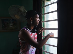 caption: Gabriel Berrio Fabré, 18, looks out the window of his room in Los Pocitos, Havana. He is a visual artist, and like most of his friends, he wants to leave Cuba once he finishes his studies.